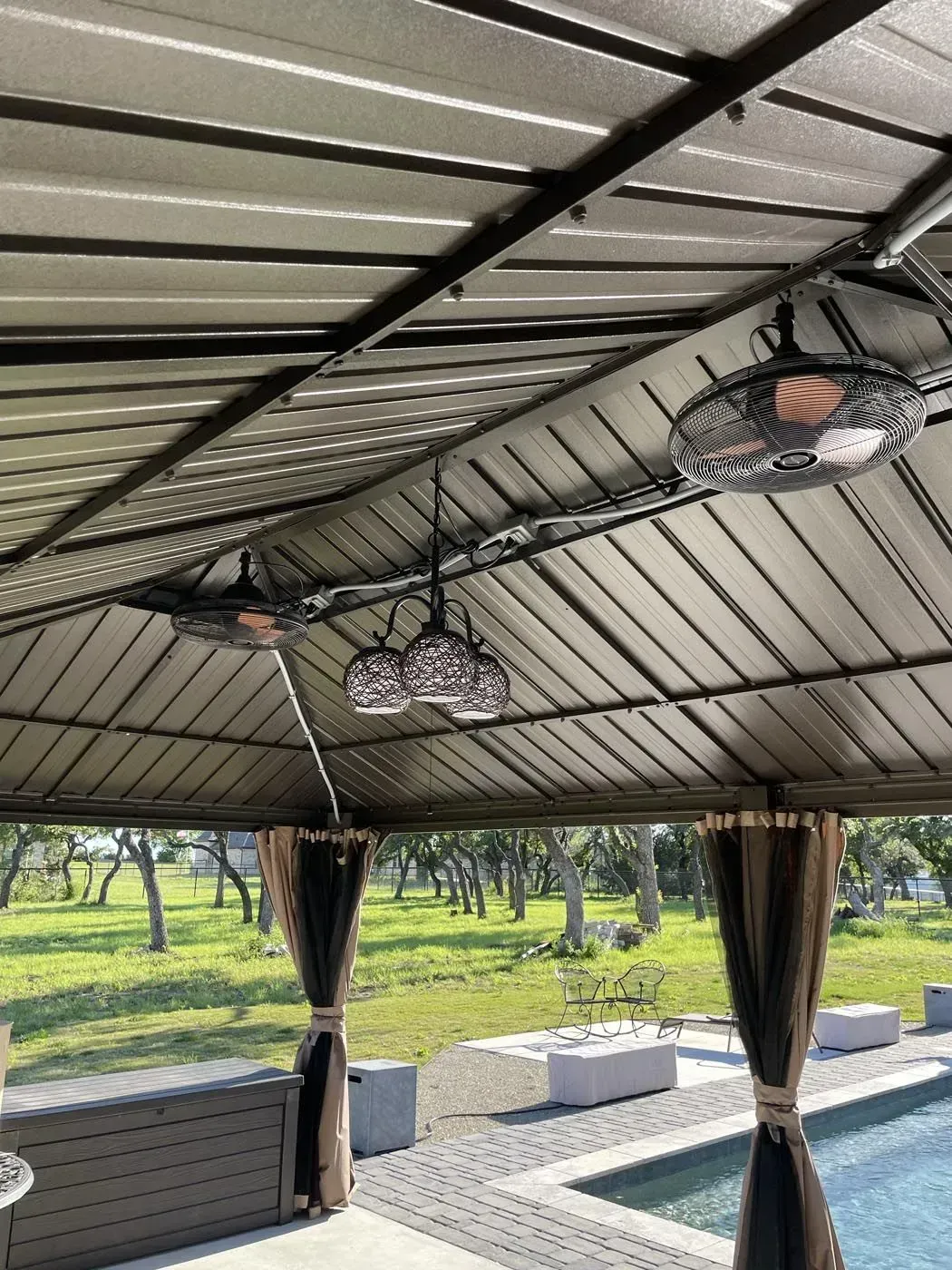 Gazebo with heaters, fans, and hanging lights over a pool. Trees and green grass in background.