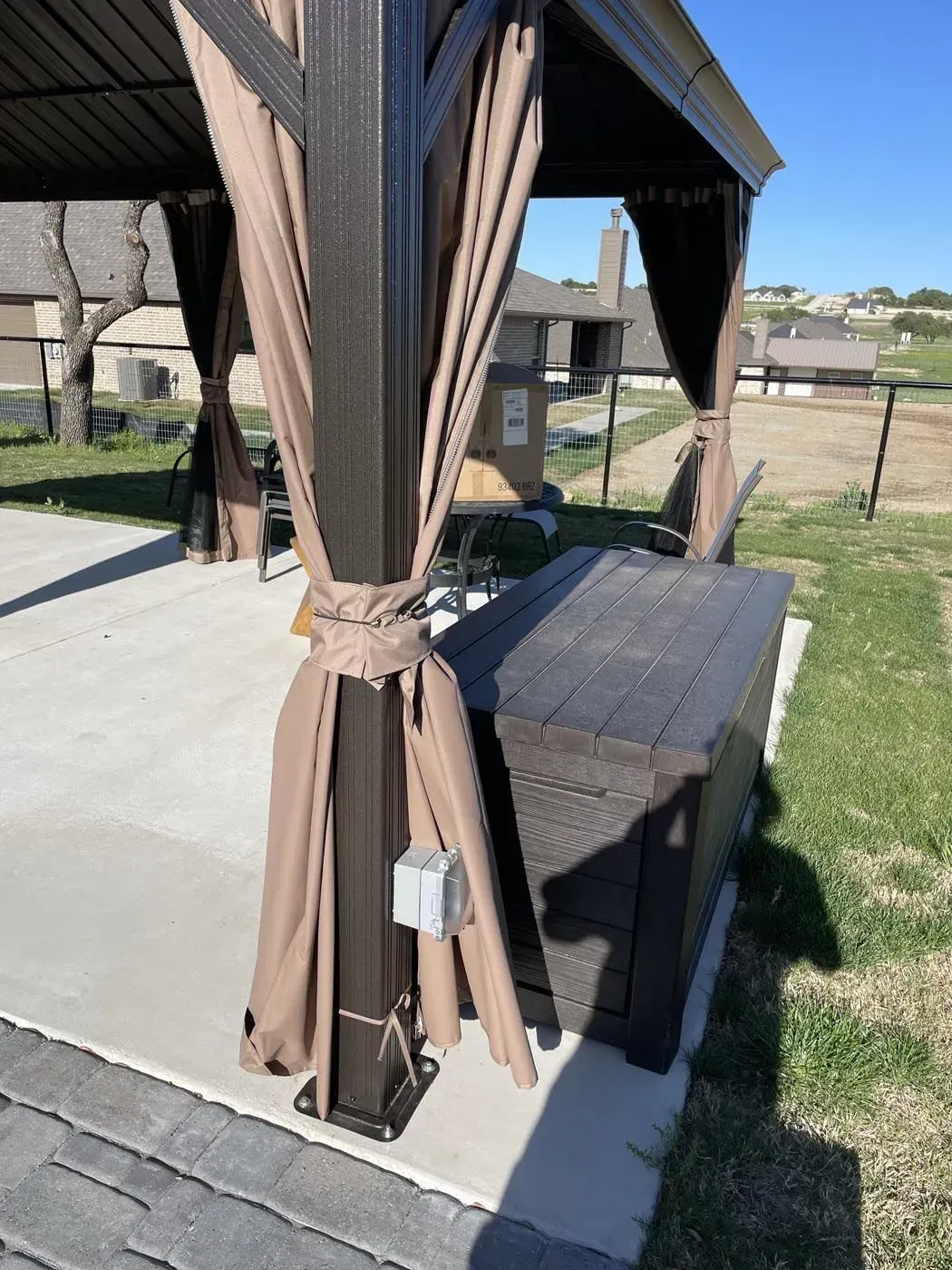 Gazebo on concrete patio with brown curtains, a storage box, and houses in the background.