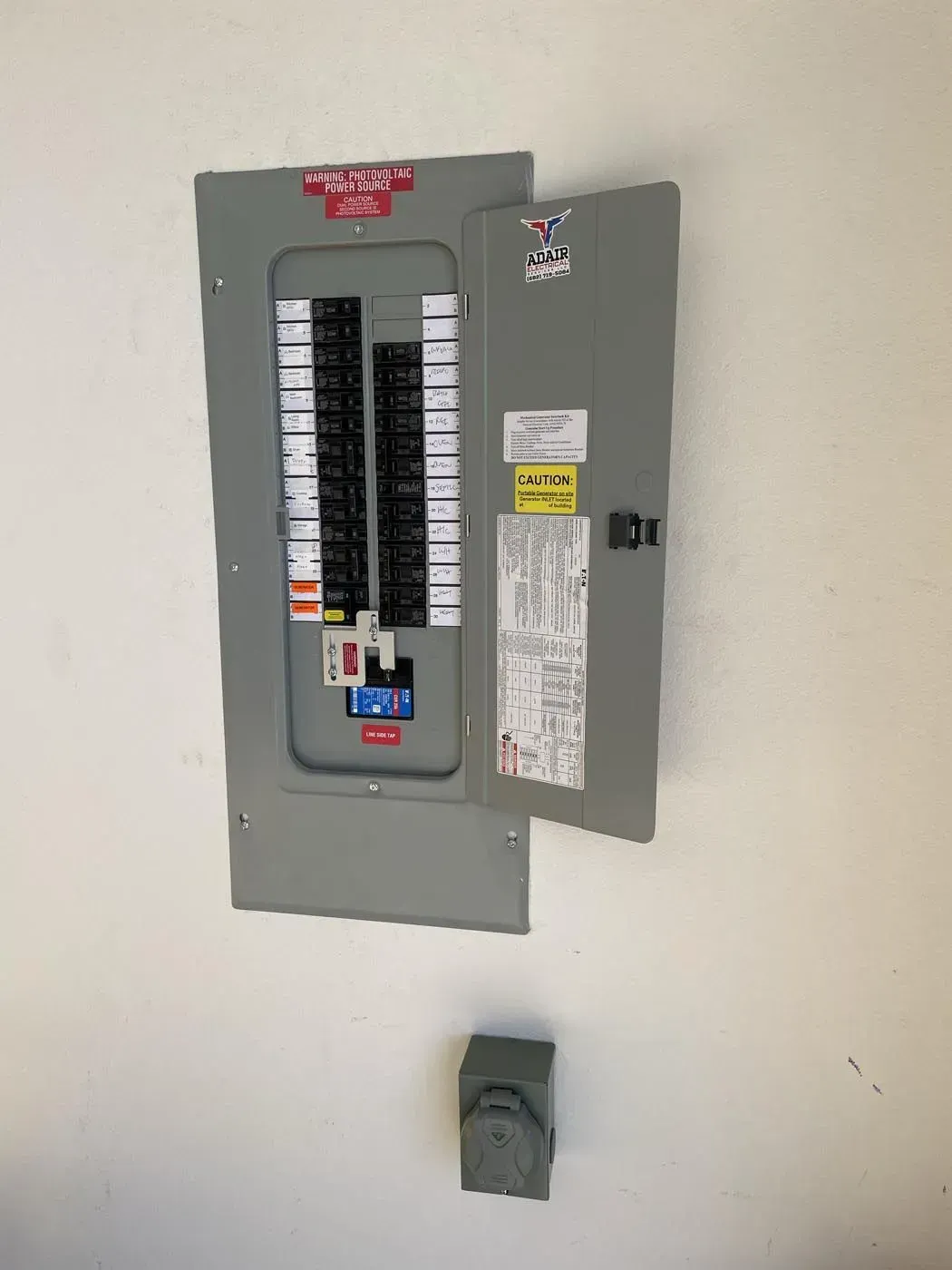 Electrical circuit breaker box mounted on a white wall, door open. A small gray box is below.