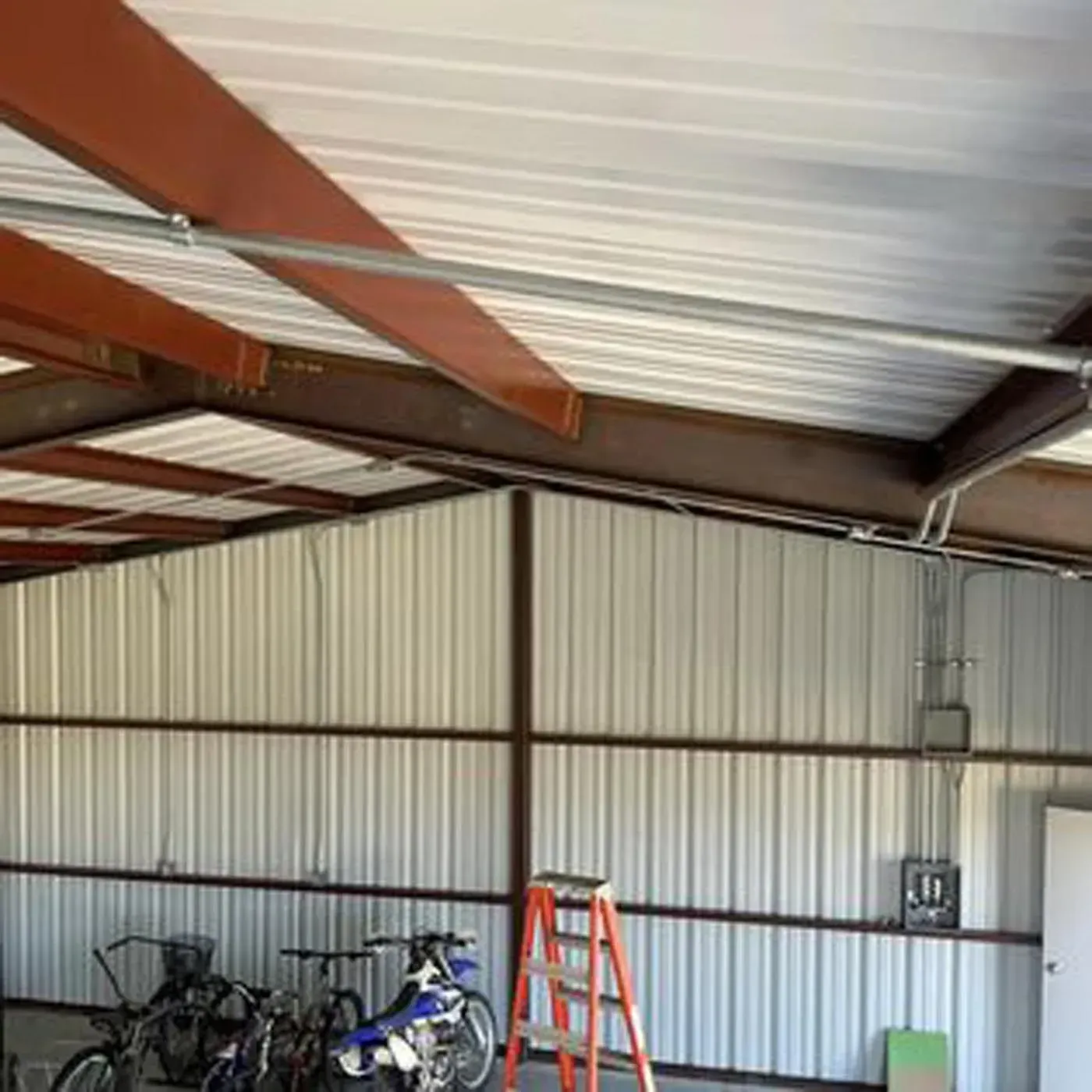 Interior of a metal building with brown beams, white corrugated metal walls and ceiling, bikes, and a ladder.