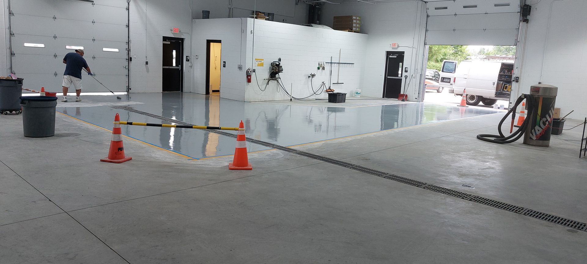 A person in a warehouse space applies a glossy epoxy coating to a section of the concrete floor marked by orange cones.