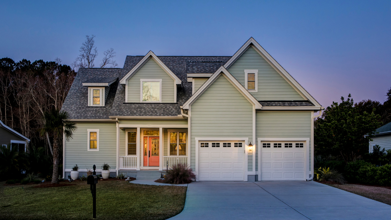 A light green, two-story house with a double garage, white trim, and a concrete driveway at twilight.