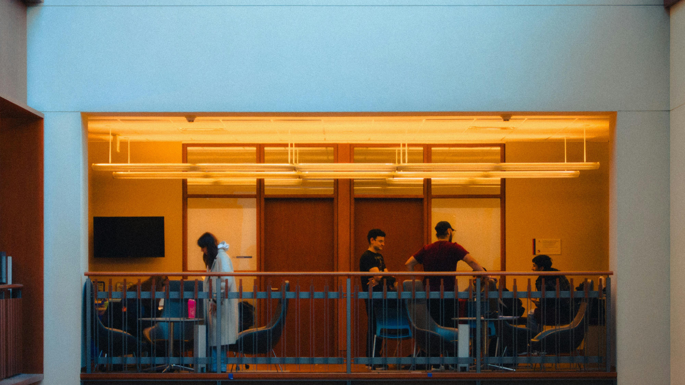 A group of people lounge and work in a warmly lit, open-plan indoor balcony area with comfortable seating.