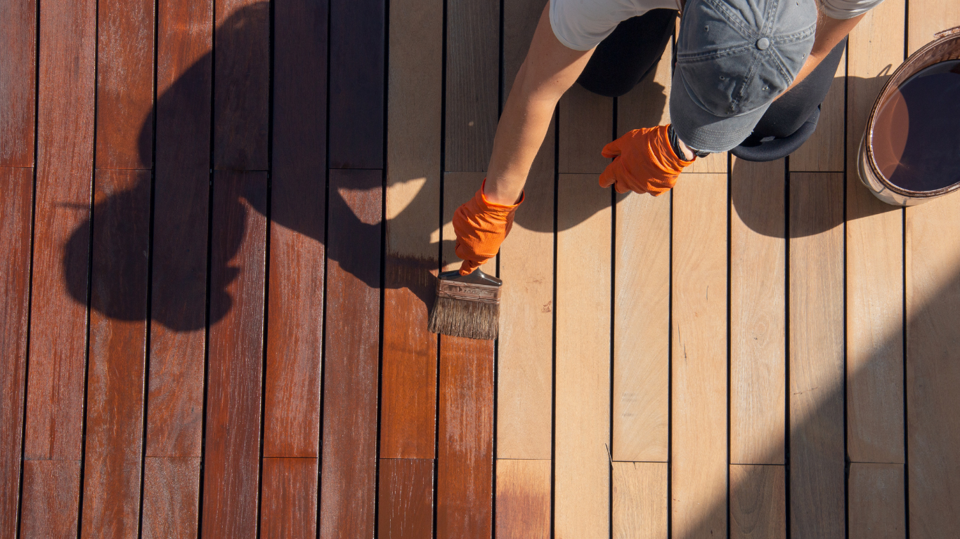 A person wearing orange gloves uses a brush to stain a wooden deck a darker, reddish-brown color.