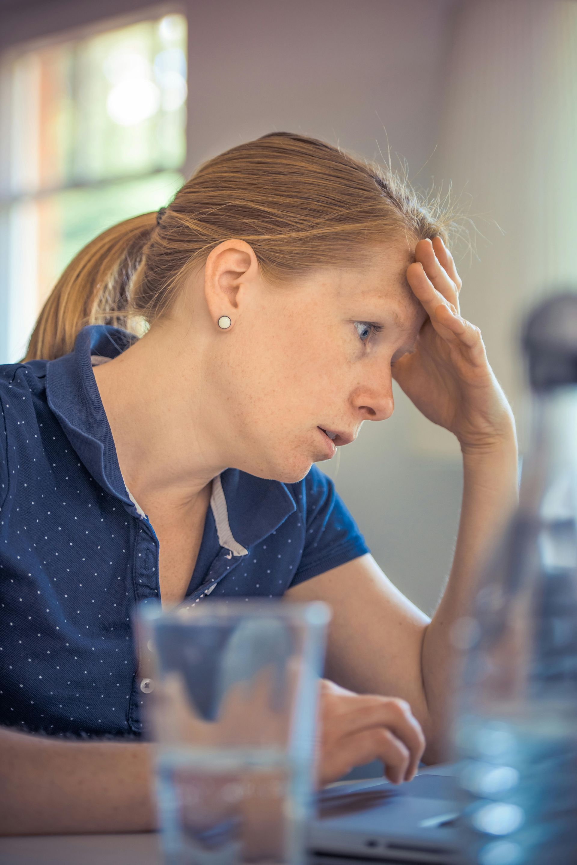 Woman with fair skin and red hair, holding her forehead, looking stressed at a laptop.