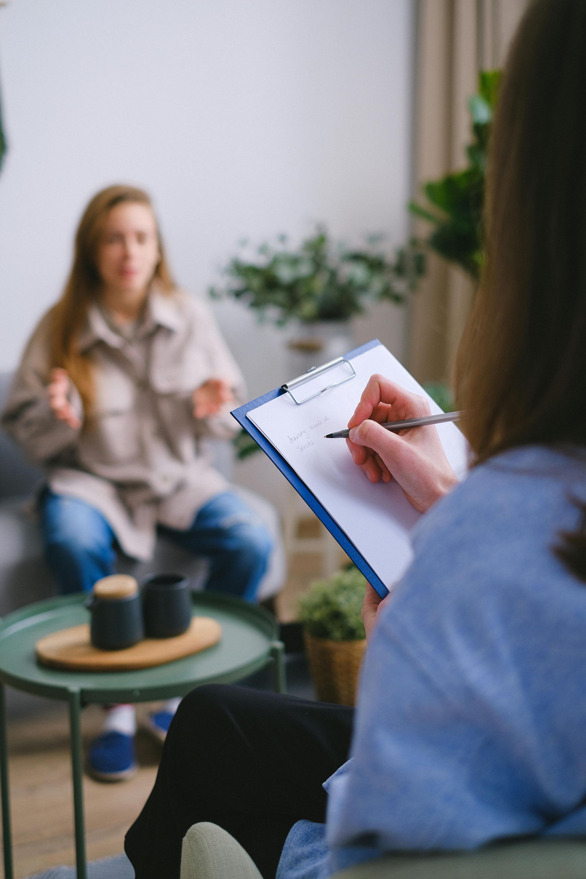 A woman is sitting on a couch talking to a female therapist while holding a clipboard.
