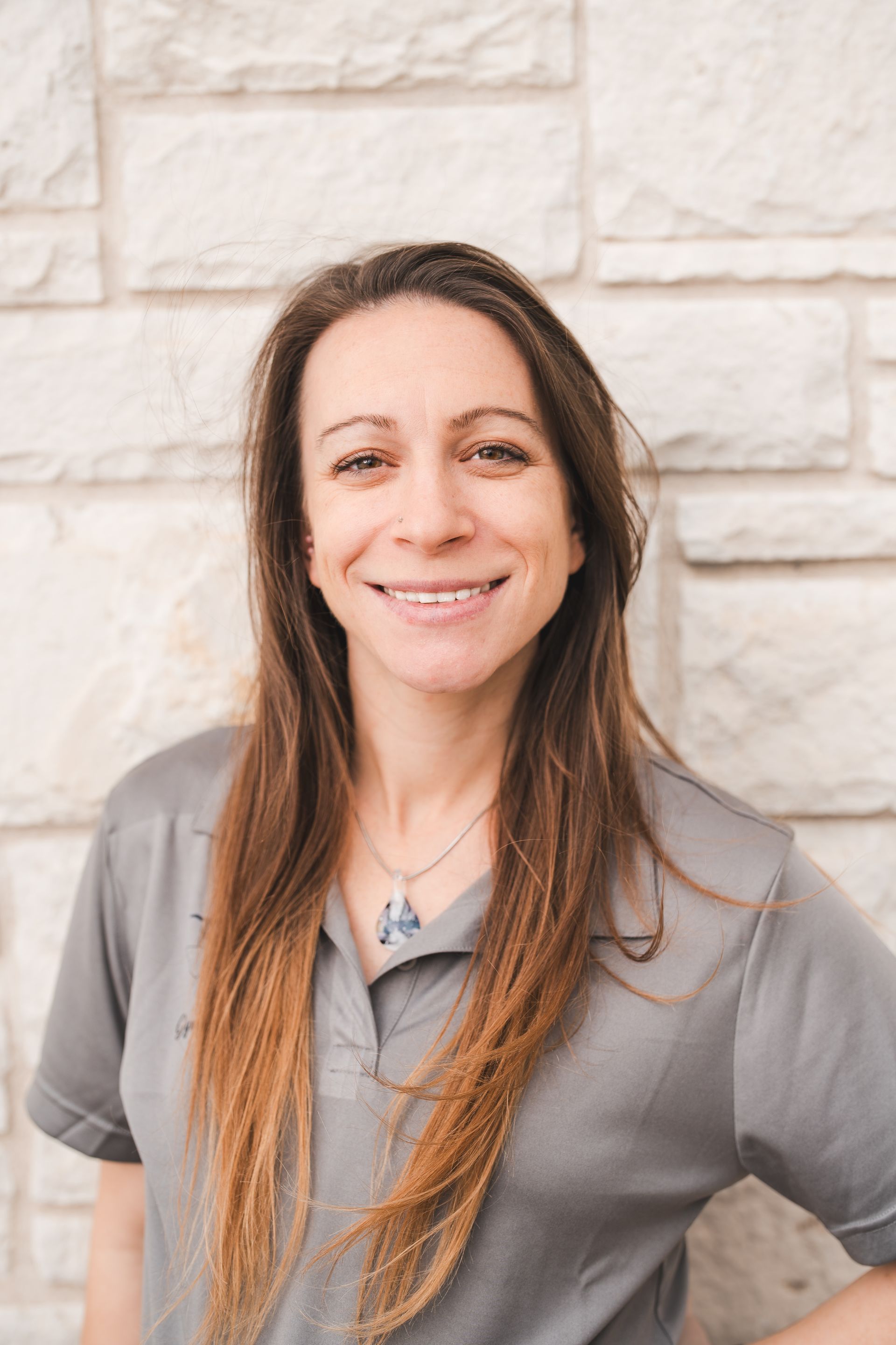 A woman in a grey shirt is smiling in front of a brick wall.