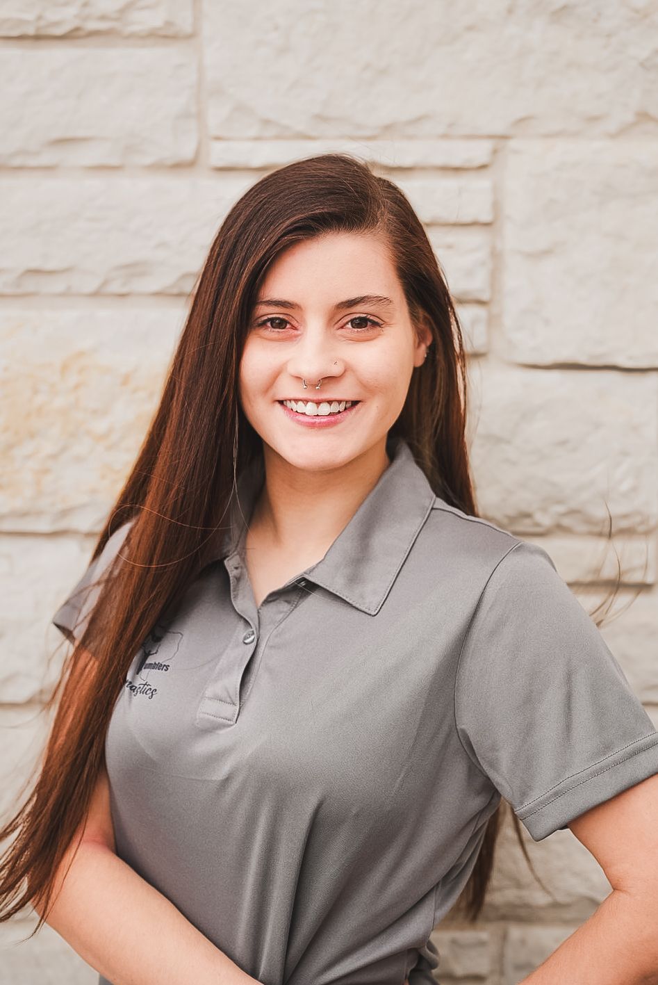 A woman with long hair is wearing a grey polo shirt and smiling.
