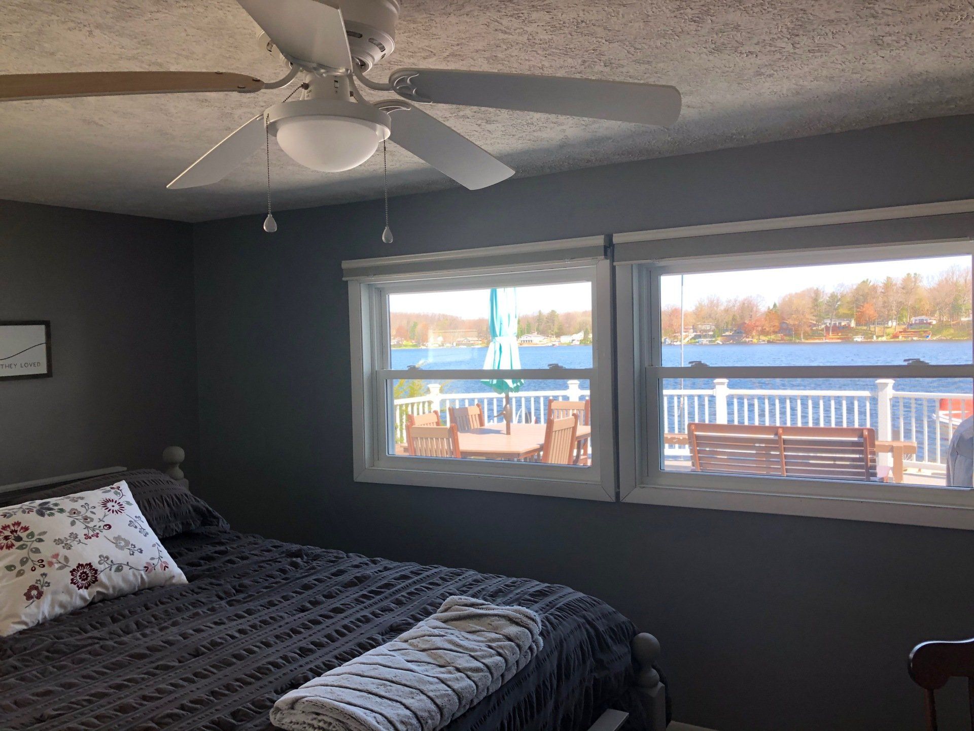 A bedroom with a bed, ceiling fan, and two windows overlooking a lake.