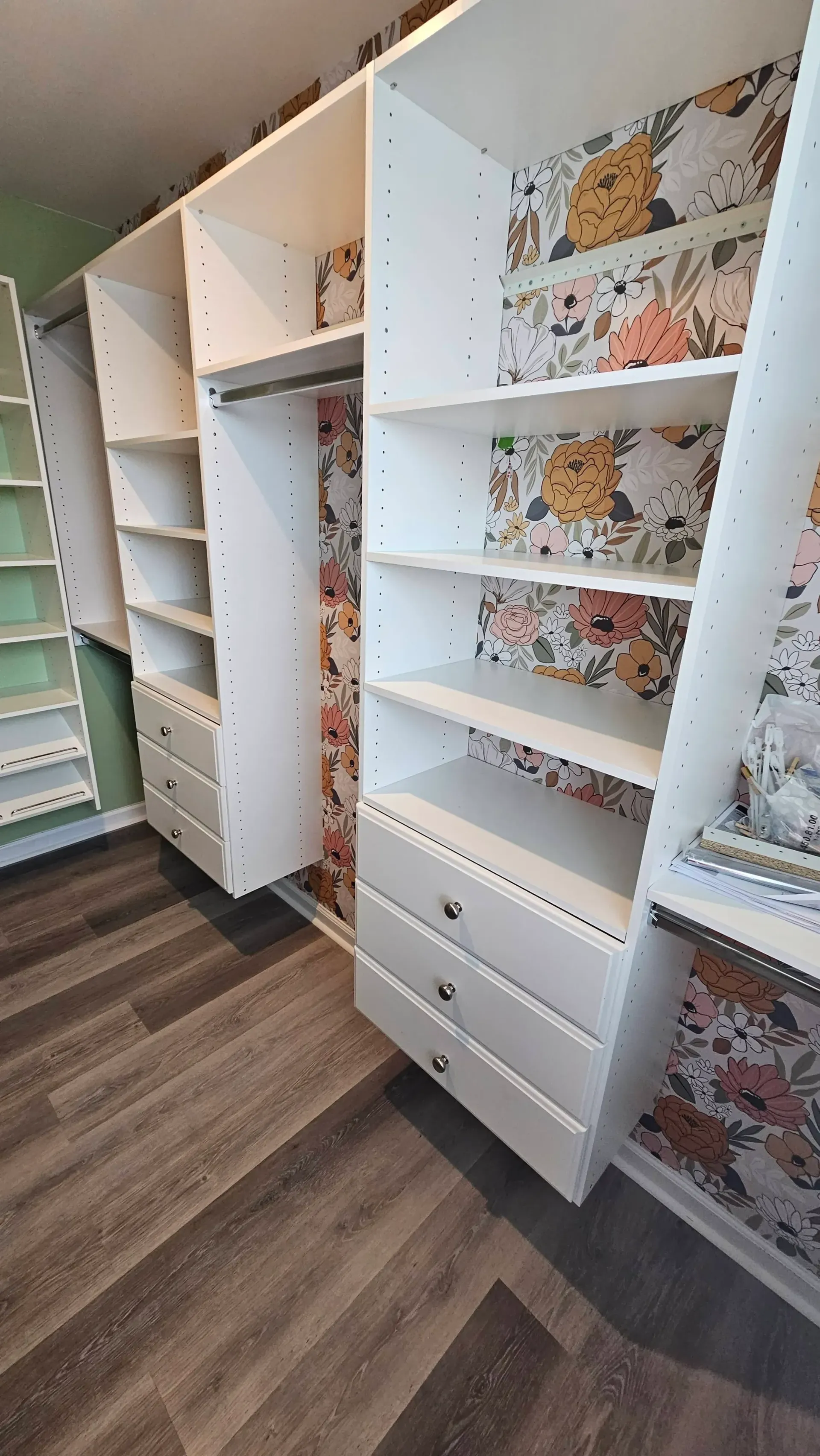 A walk in closet with white shelves and drawers and floral wallpaper.