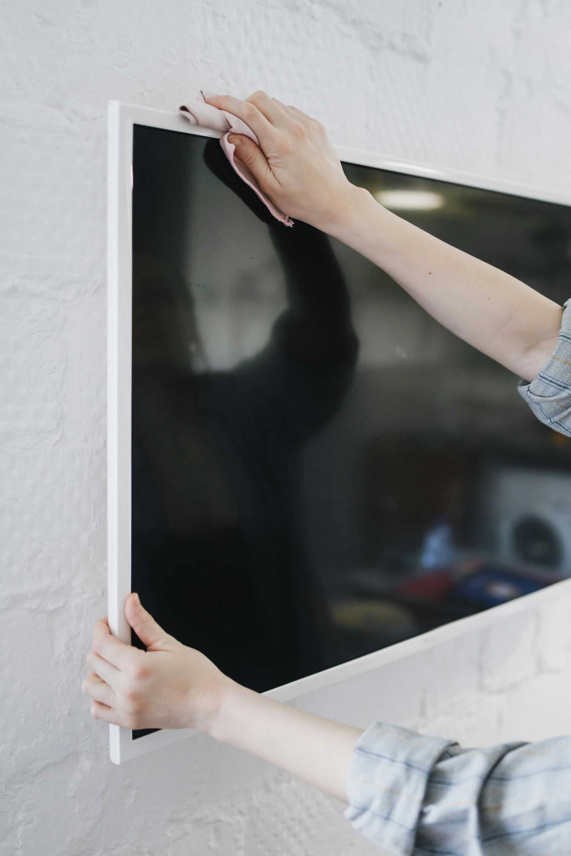 A person cleaning the frame of a wall-mounted flat-screen television with a white cloth.