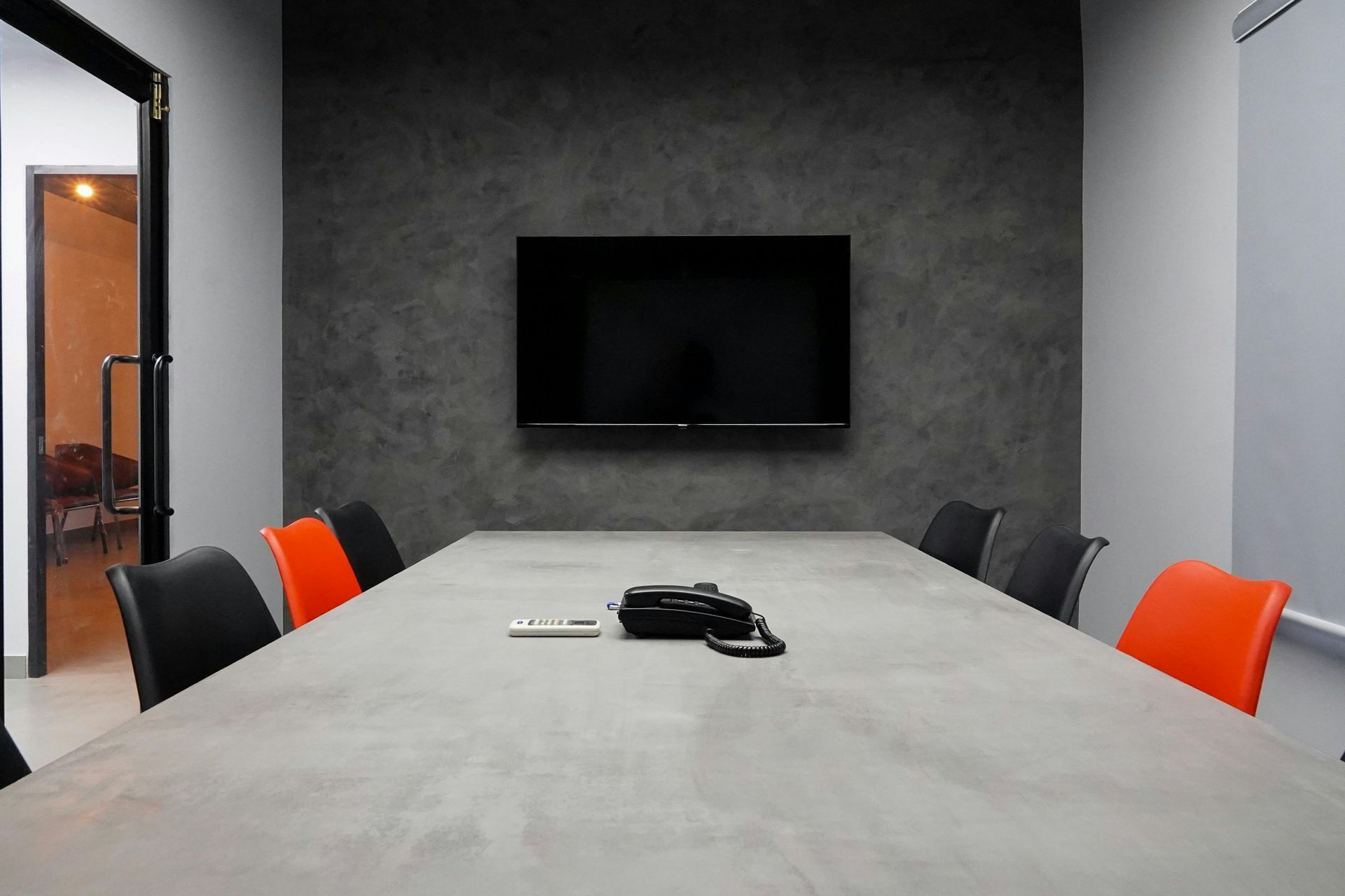 A modern meeting room features a concrete-style table, black chairs, orange accents, and a wall-mounted TV.