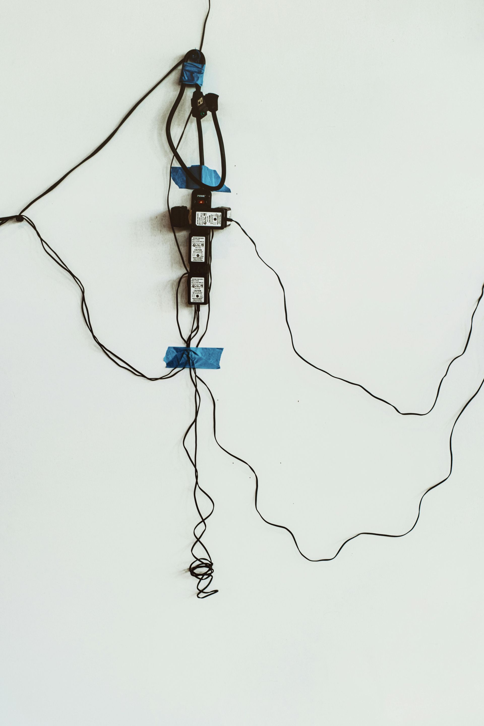 A vertical power strip attached to a white wall with blue painter's tape, with several tangled black cords hanging from it.
