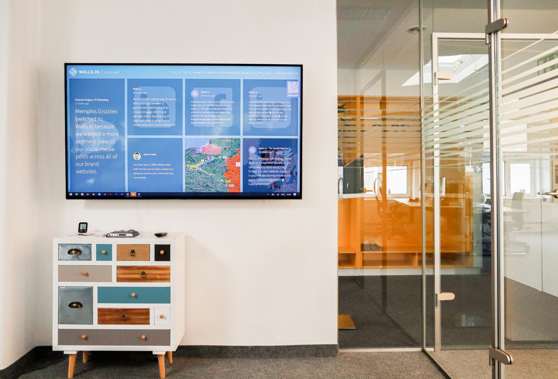 A wall-mounted display showing a digital dashboard hangs above a small, multi-colored chest of drawers in a modern office.
