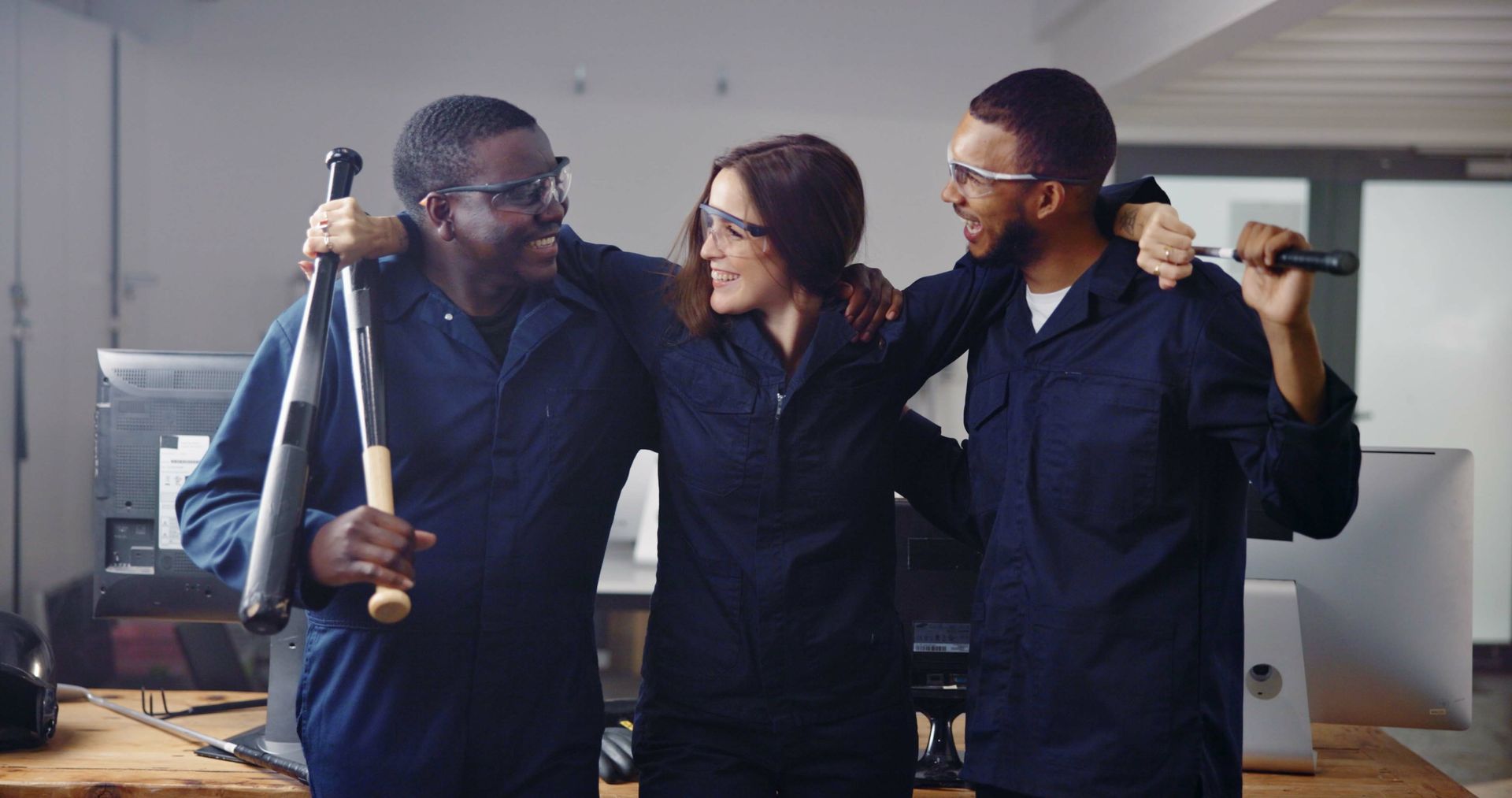 Three people in blue coveralls pose with baseball bat and wrench in office setting.