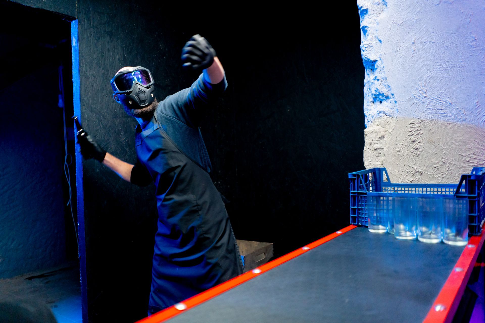 Person in mask and gloves reaching, near a tray of glasses and a doorway.