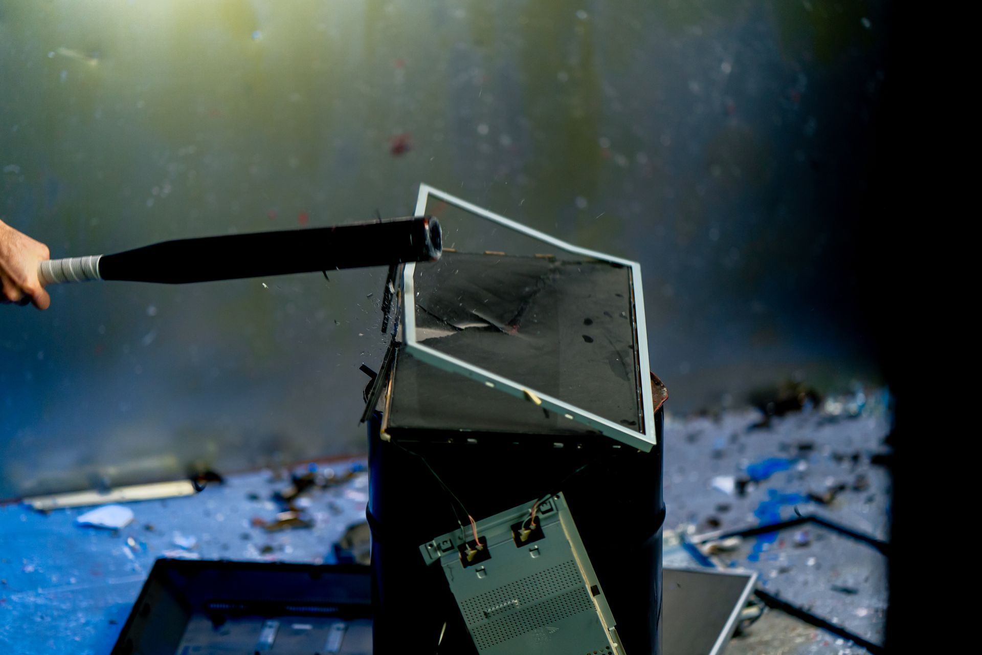 A person's arm swings a black bat, smashing a computer monitor. Blue room.