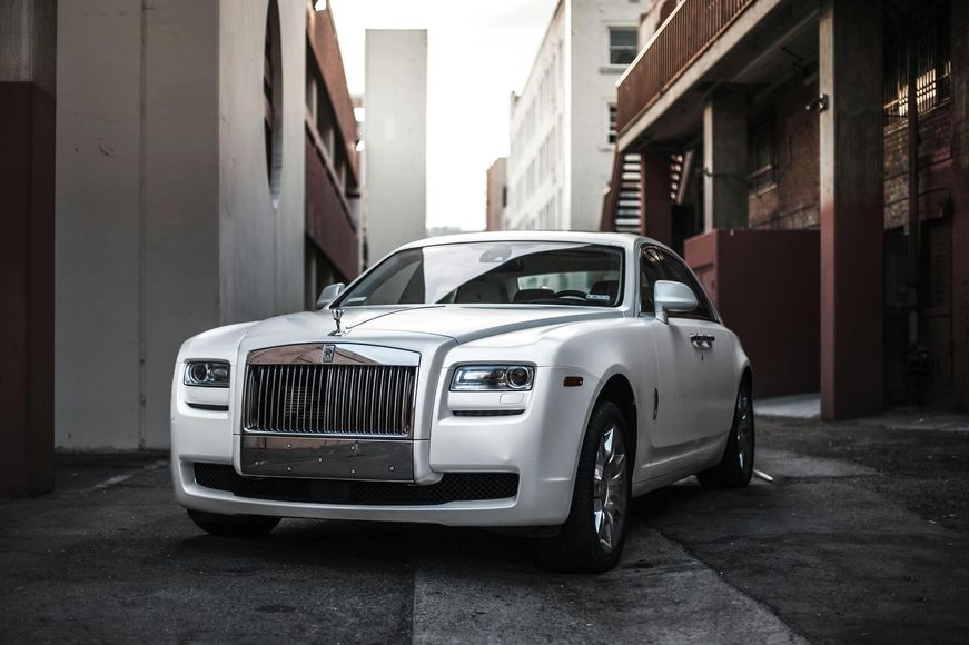 A white rolls royce is parked in a parking lot between two buildings.