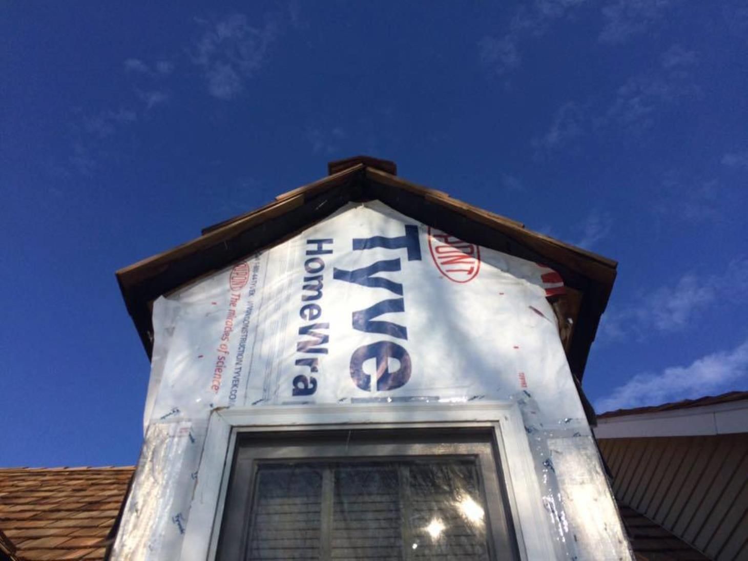 A house under construction, with Tyvek HomeWrap covering the exterior, a window, and a brown roof against a blue sky.