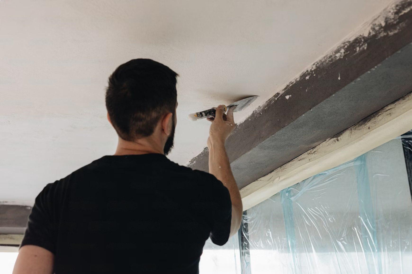 Person on a ladder using a putty knife to smooth plaster on a ceiling. Dark trim and plastic-covered window visible.