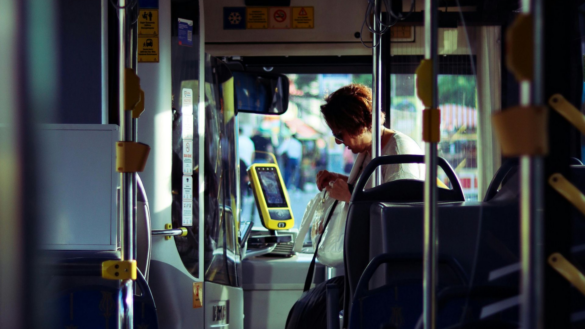 Inside a bus, person seated near a ticket validation machine, view from the aisle.