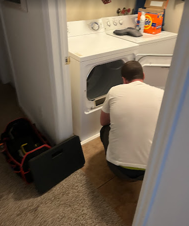 A man is kneeling down in a laundry room next to a washer and dryer.
