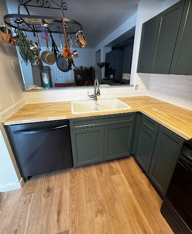 A kitchen with green cabinets , a sink , a dishwasher , and a wooden counter top.