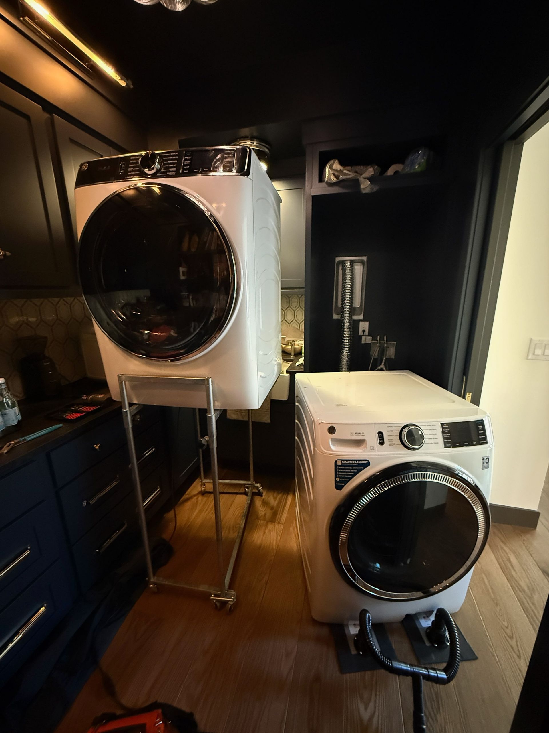 A white washer and dryer in a dark-themed laundry room, with one appliance elevated on a metal stand.