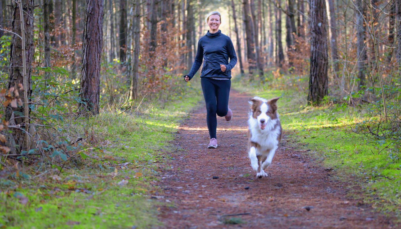 A woman is running with her dog on a trail in the woods.