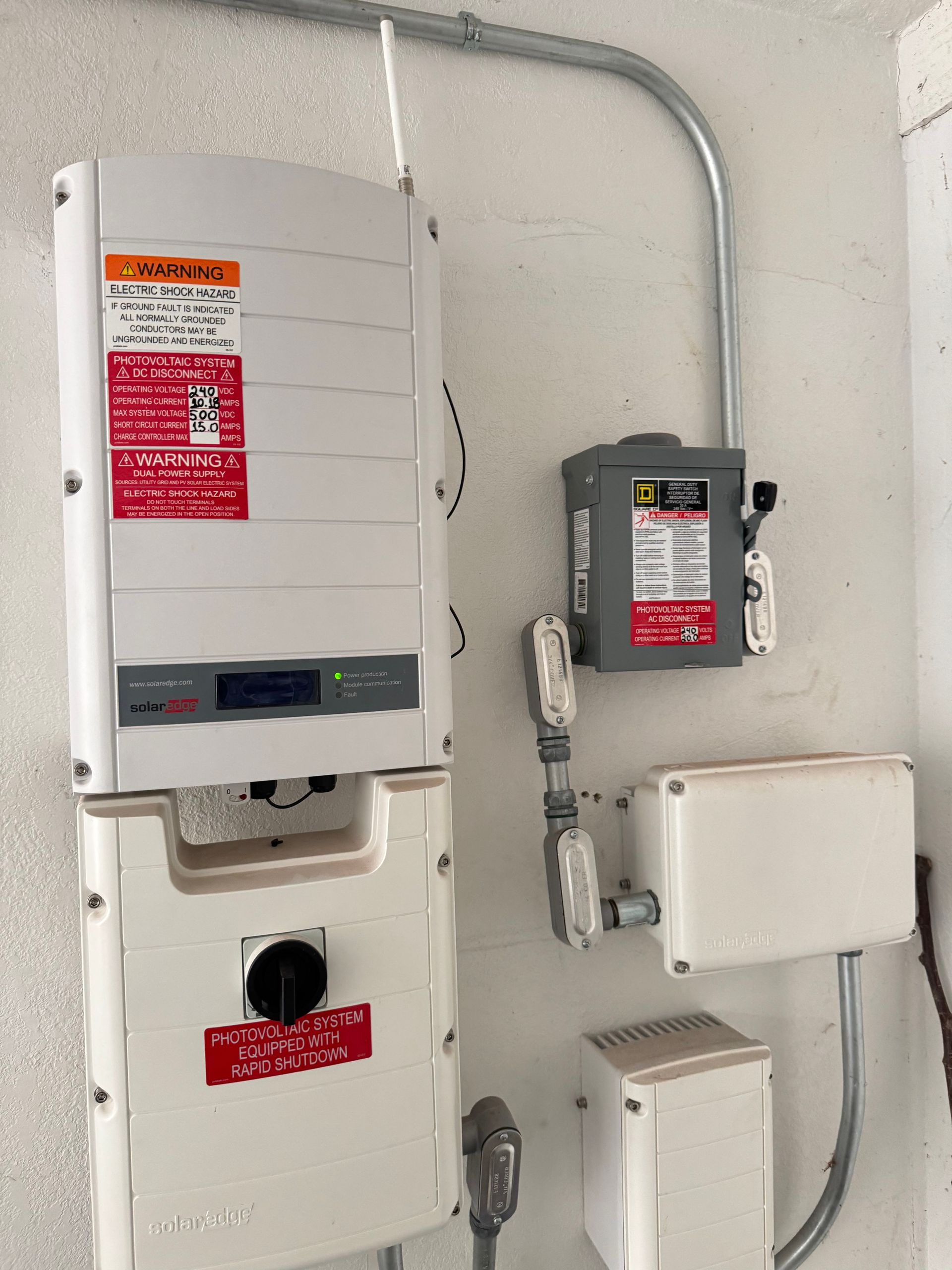 Wall-mounted solar power inverter with adjacent gray metal disconnect switch and utility junction boxes on a white wall.