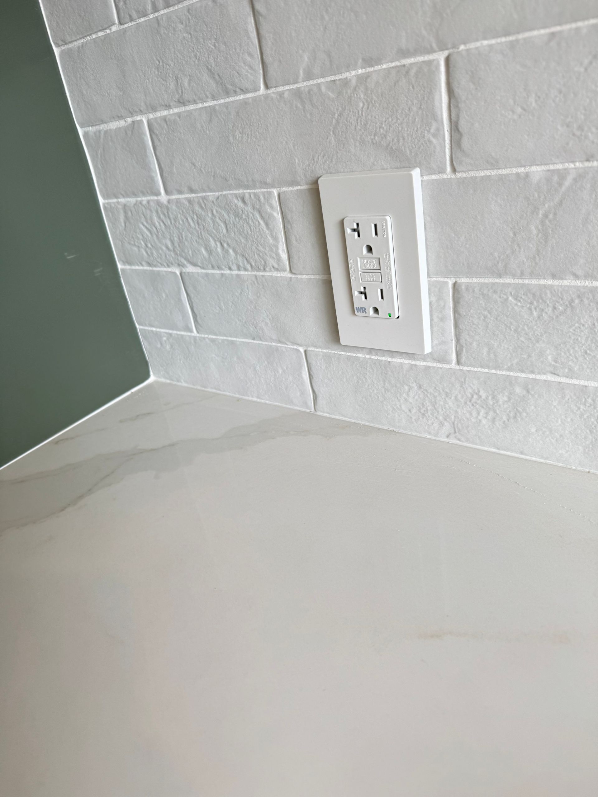 A white GFI wall outlet mounted on a textured, light gray brick backsplash above a white countertop.