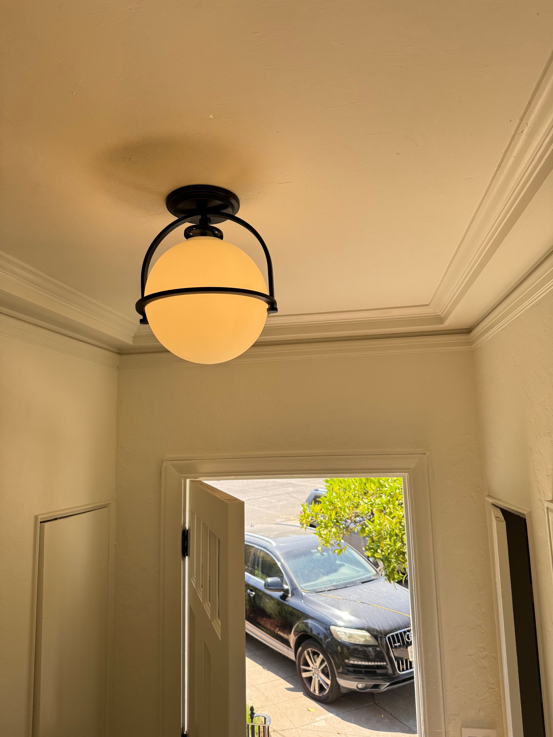 A black spherical ceiling light fixture hanging in an entryway, with an open door showing a car parked outside.