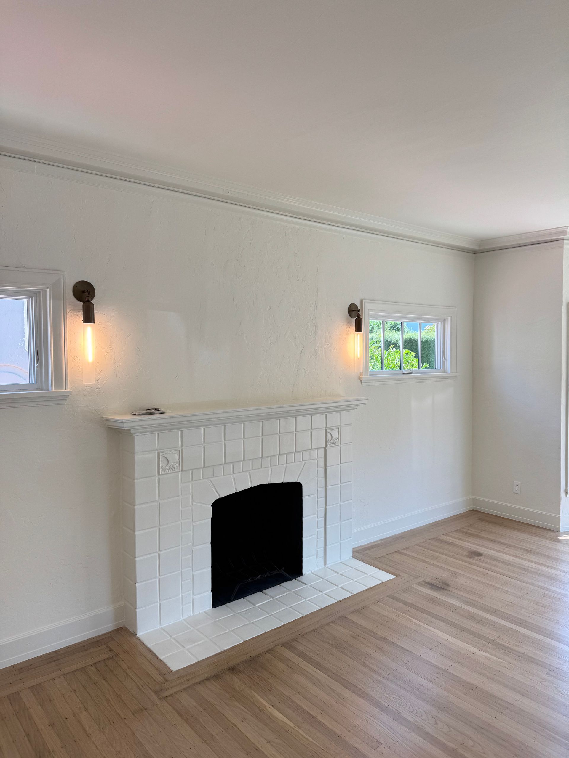 A bright, empty living room with a white brick fireplace, hardwood floors, and two wall-mounted light fixtures.