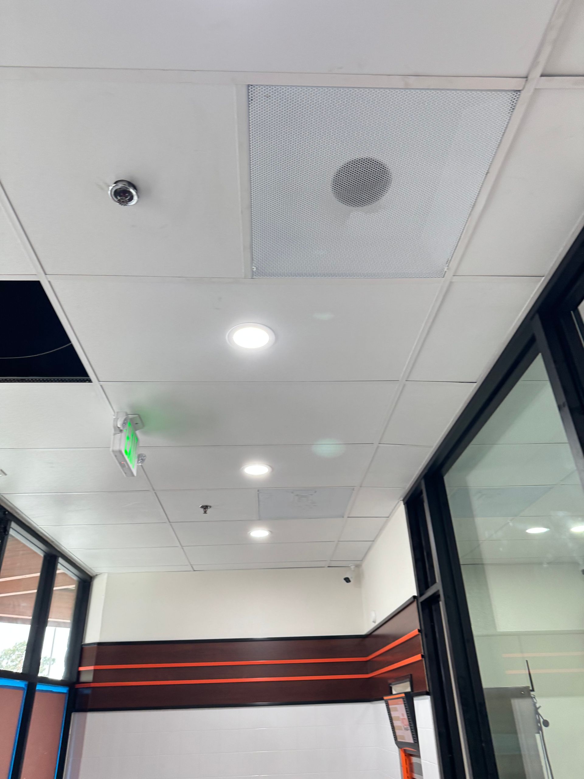 A low-angle view of a drop ceiling with recessed lighting, a ventilation grate, a camera, and an exit sign in a hallway.