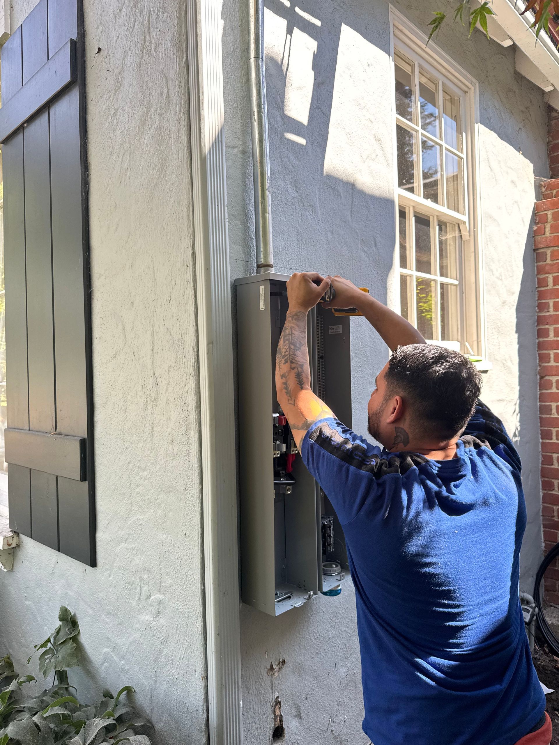 A person in a blue t-shirt works on an electrical meter box attached to the exterior wall of a building.
