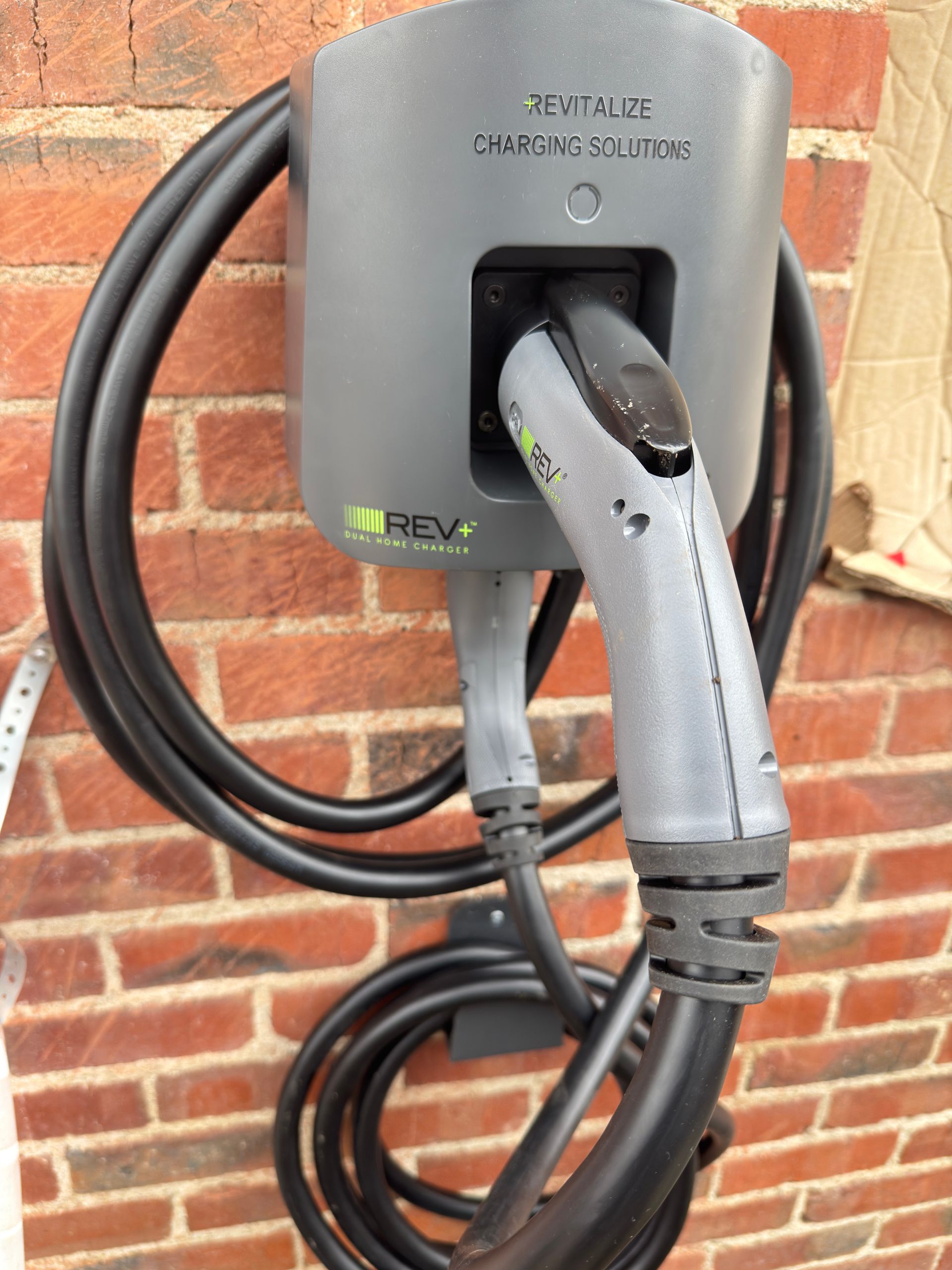 A wall-mounted grey REV charging station unit with a black charging cable coiled underneath on a brick wall.