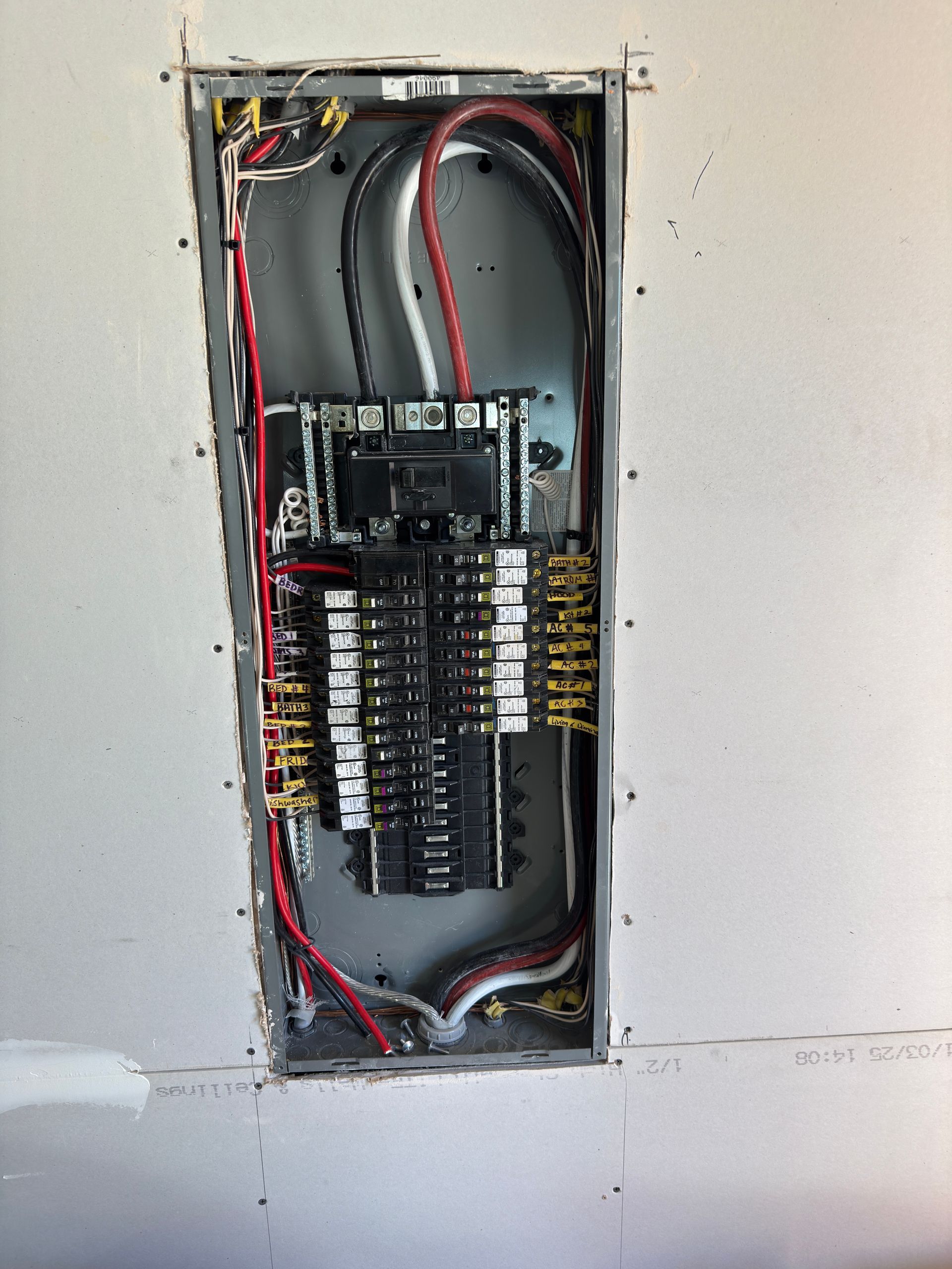 An open electrical breaker panel mounted in a drywall wall, showing interior wiring, circuits, and components.