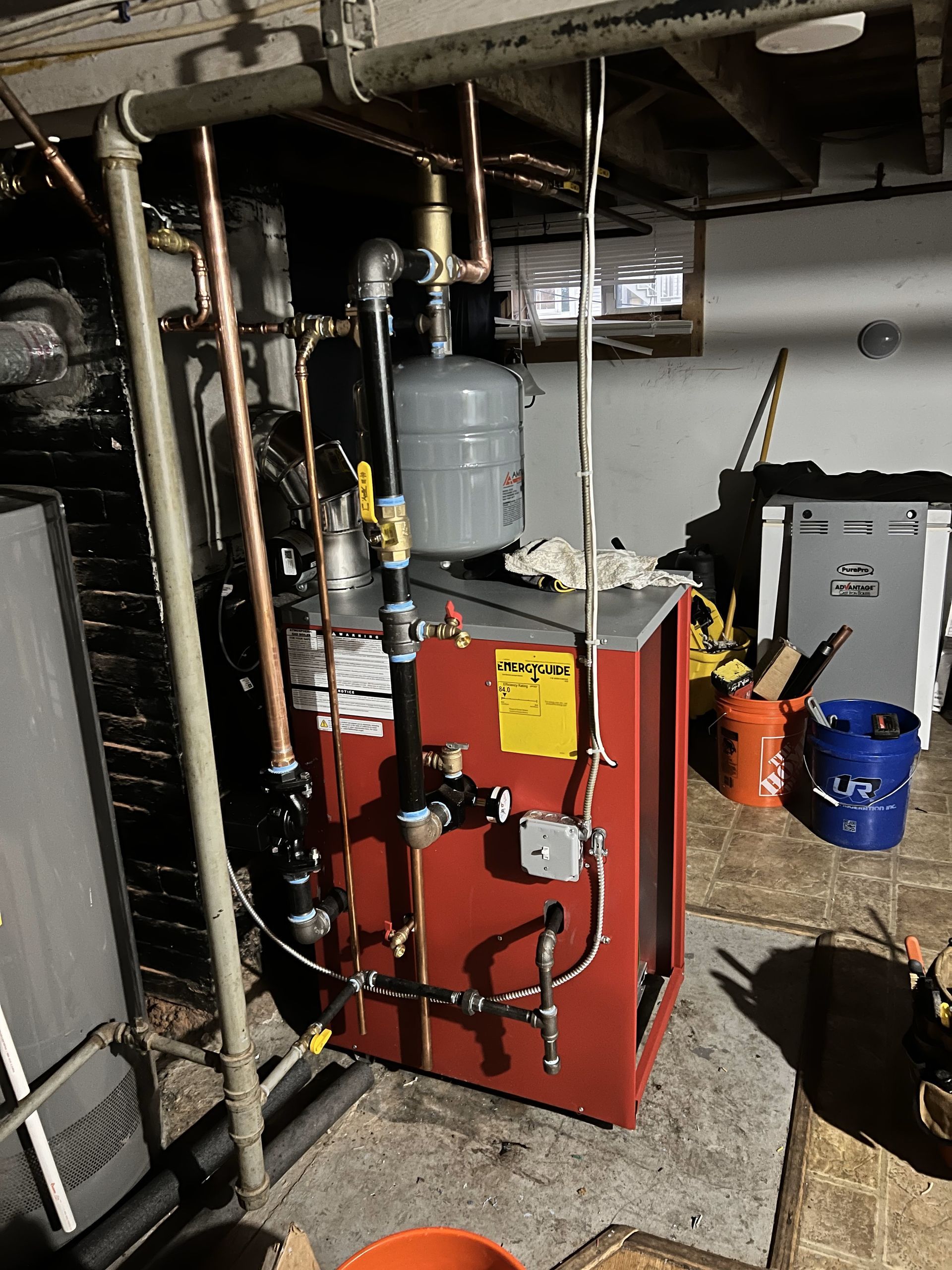 A red boiler is sitting in a basement surrounded by pipes.