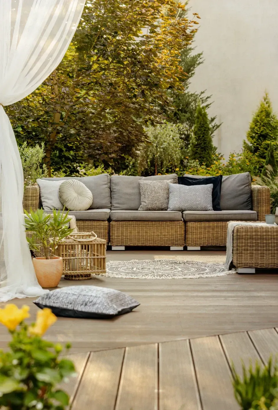 Cozy outdoor patio with beige sectional sofa, wicker table, cushions, and potted greenery under soft curtains