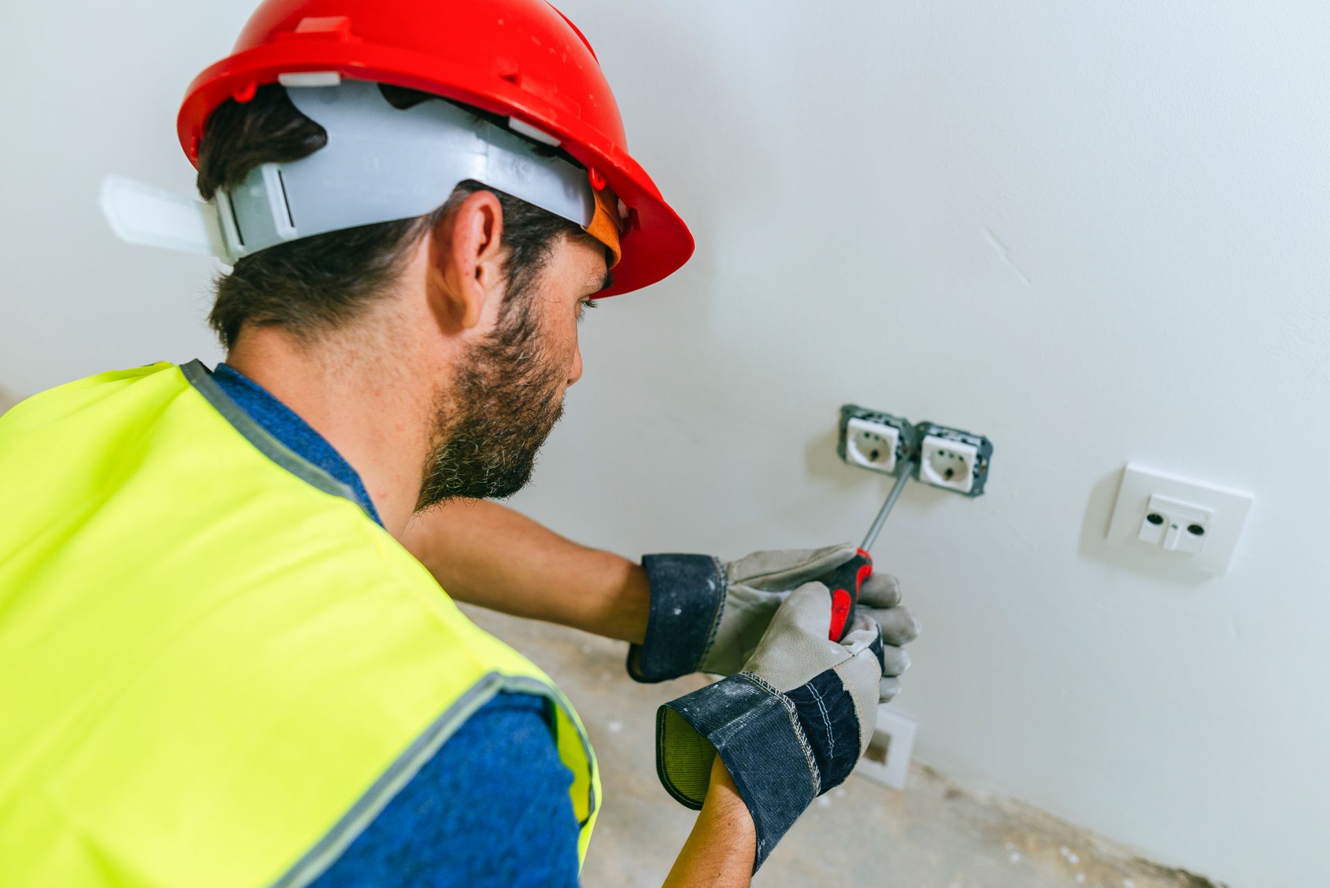 A man wearing a hard hat and safety vest is working on an electrical outlet.