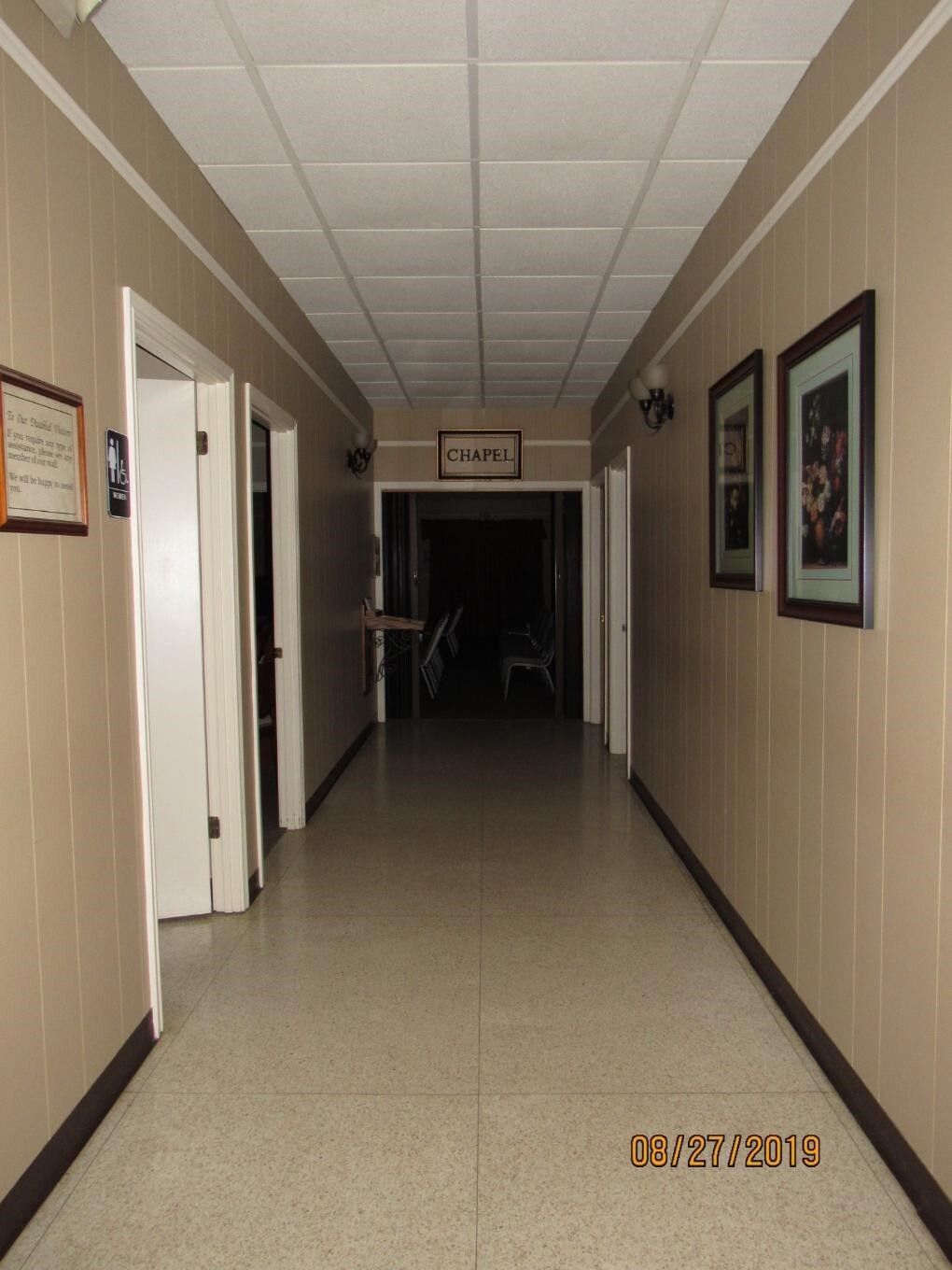 Long, beige hallway with doors on either side, leading to a dark room at the end; framed art on the walls.
