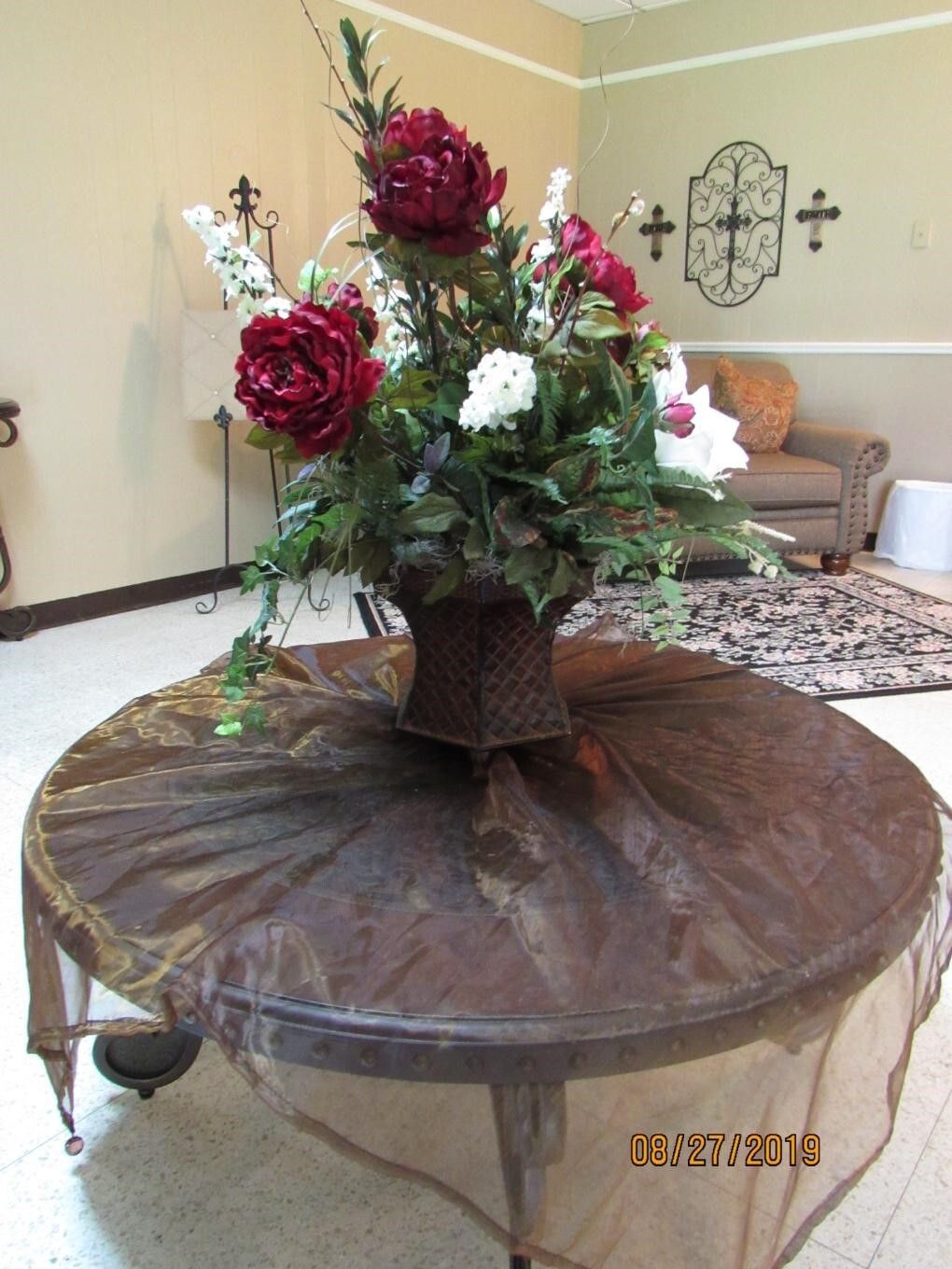 Large floral arrangement on a round table draped with sheer brown fabric; beige wall with decorative accents.