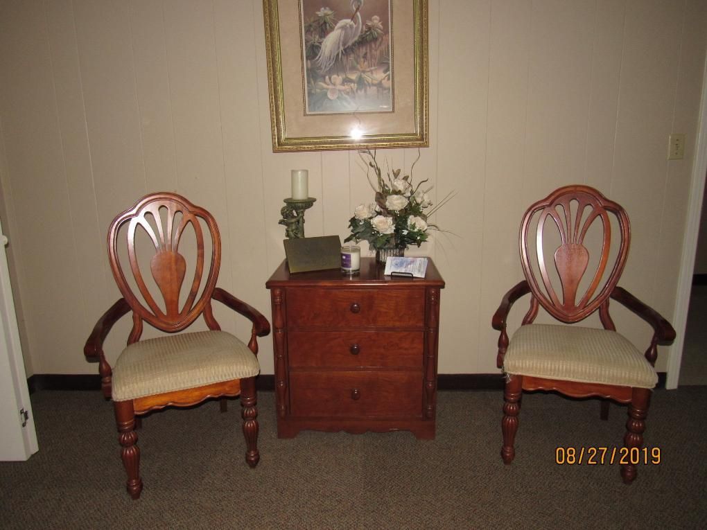 Two ornate wooden chairs flanking a wooden cabinet with a painting above; cream walls.