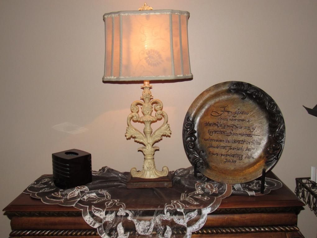 Lamp, decorative plate, tissue box on a wooden table with a lace doily, set against a beige wall.