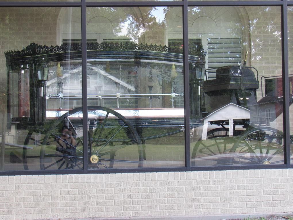 Horse-drawn hearse in a glass display case against a brick wall.