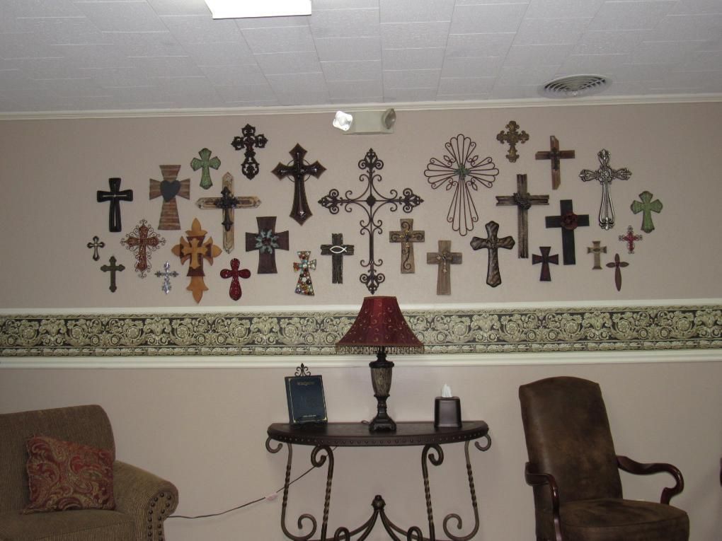 Wall with many crosses, table with lamp and items, two chairs in a room.
