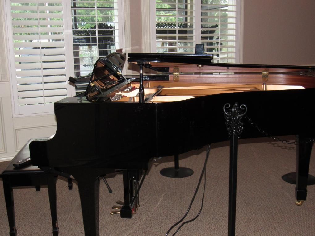 Black grand piano, open, in front of a window with white shutters, recording equipment set up.