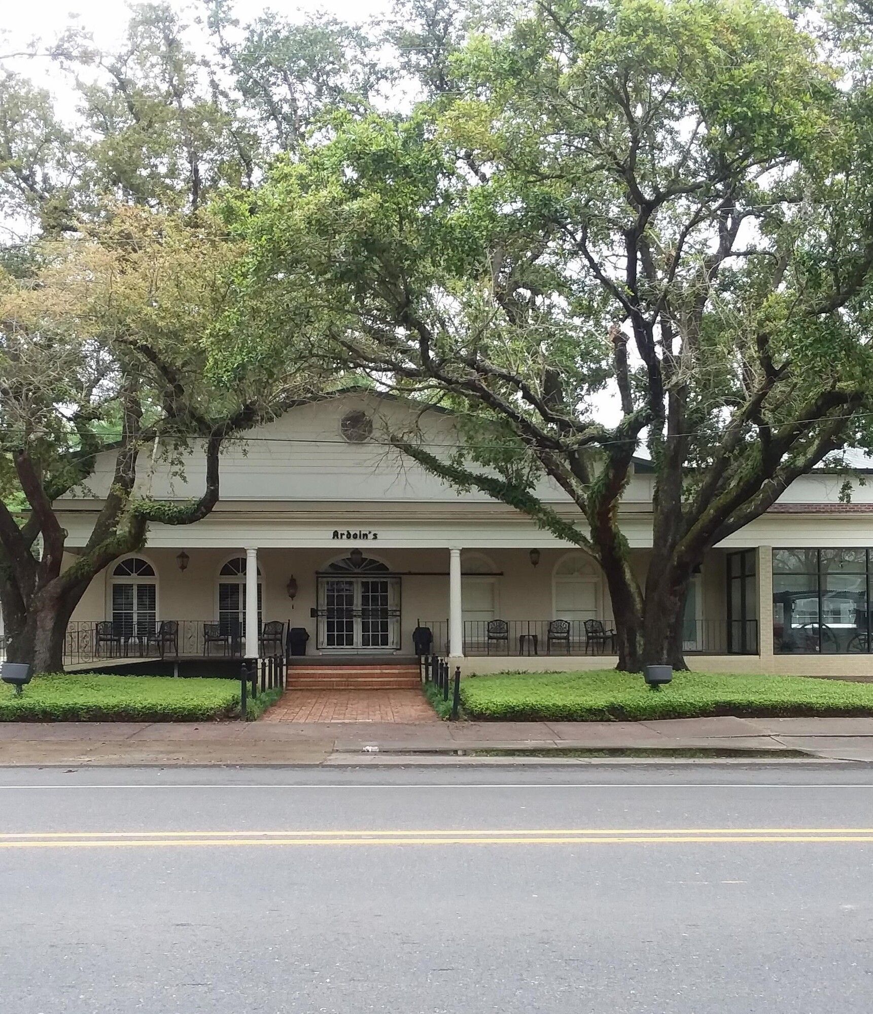 White building with porch, flanked by trees; brick walkway leads to entrance on a street.