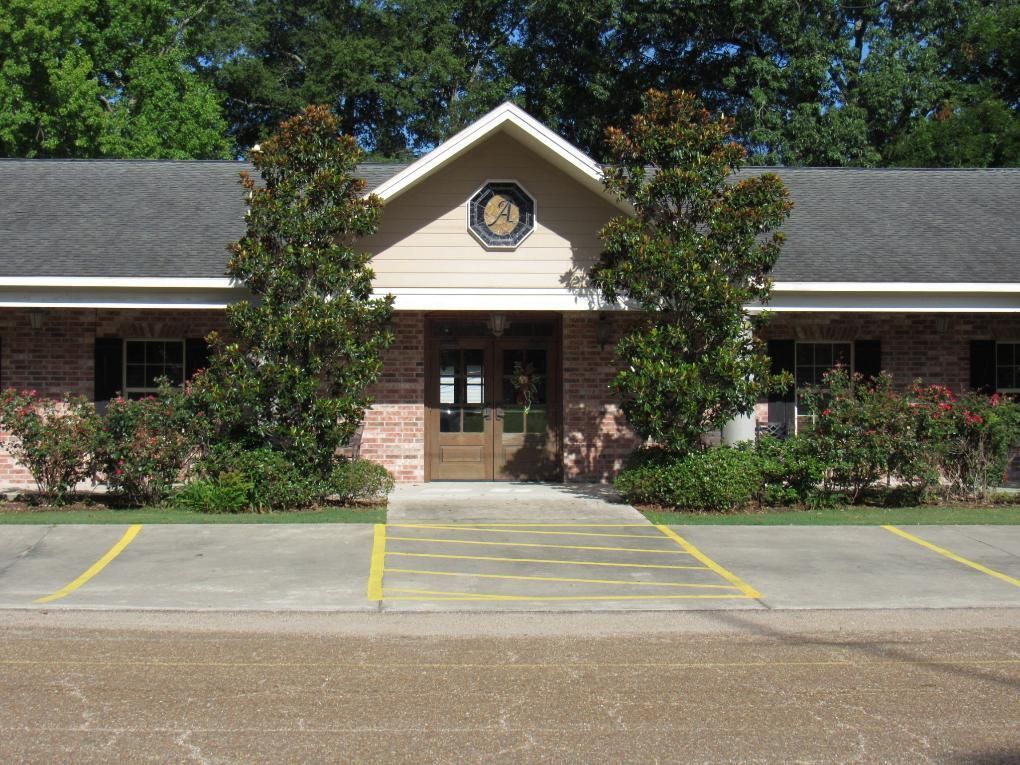 Building entrance with trees, bushes, and parking spaces, beige and brick facade, dark roof, sunny day.