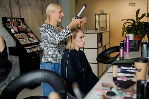 Una donna bionda arriccia i capelli di una cliente in un salone. La cliente è seduta su una sedia e indossa una mantella nera. Sullo sfondo, un espositore per il trucco e i prodotti.